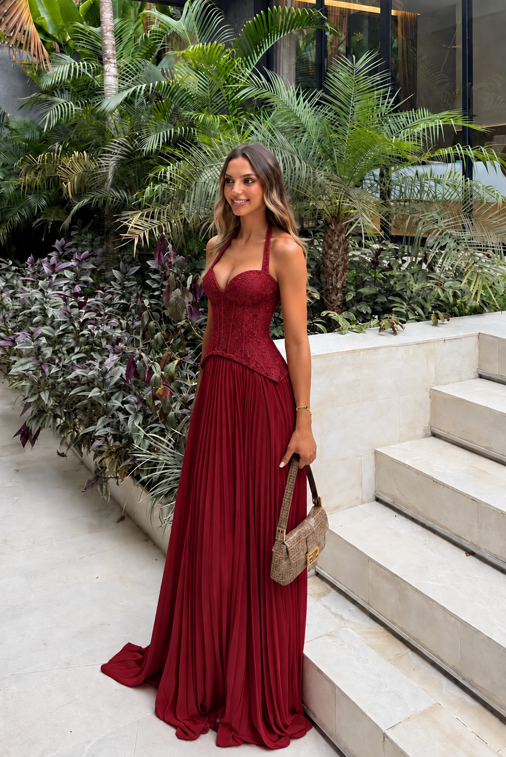 Enid Pleated Corset Maxi Dress – Wine Red worn beside tropical greenery and pale stone steps